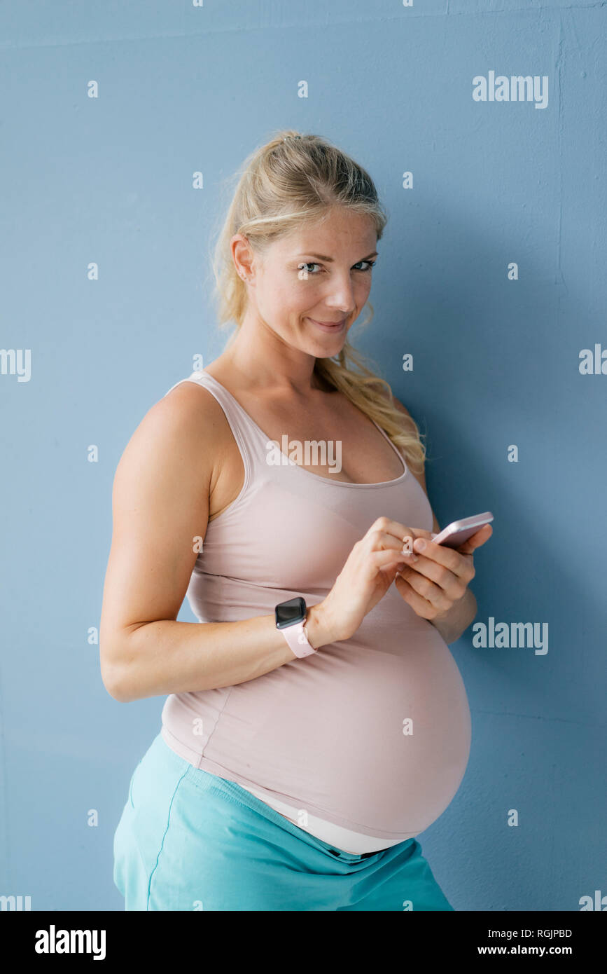 Portrait of smiling pregnant woman standing at blue wall using cell phone Stock Photo