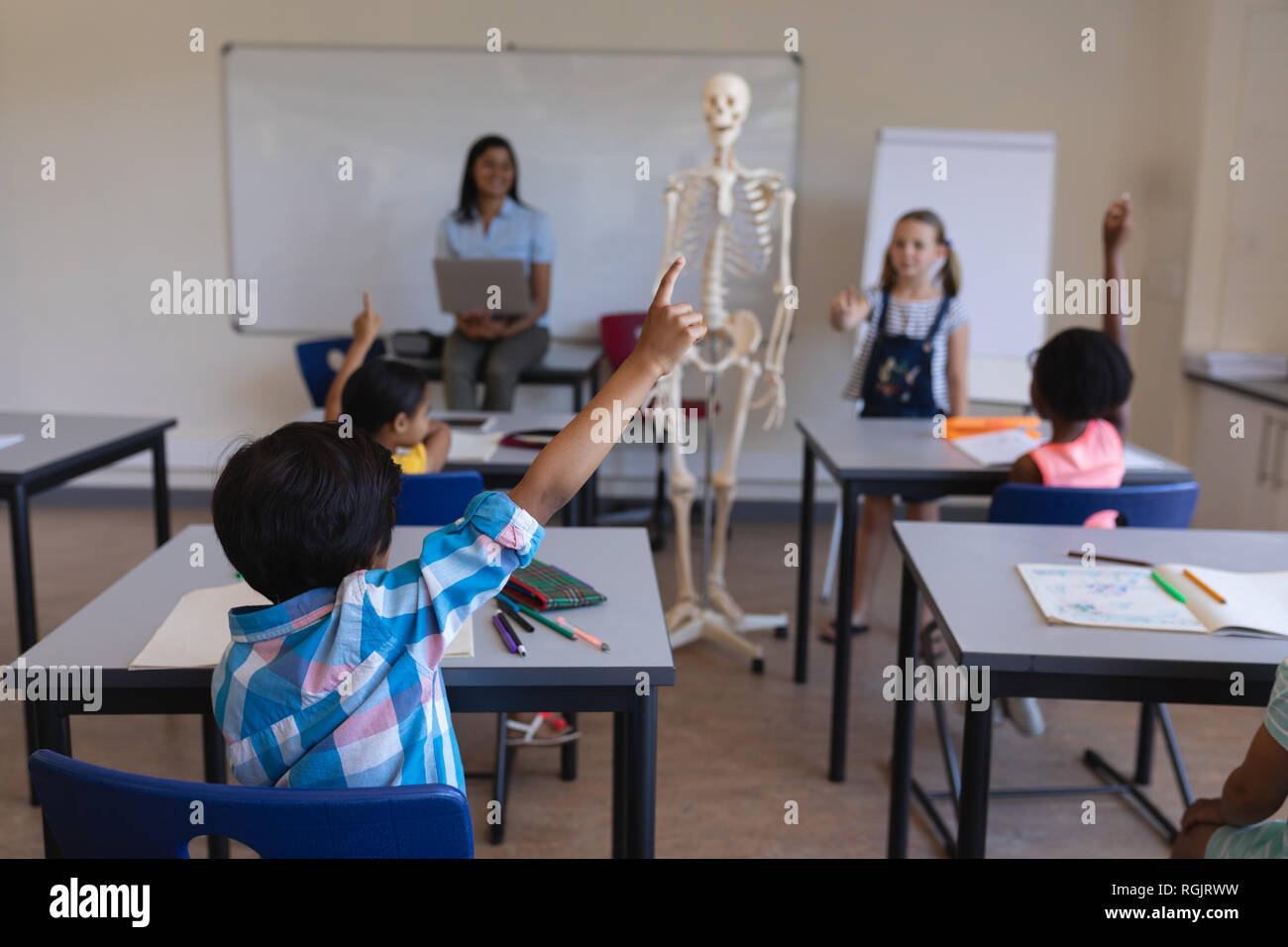 Rear view of schoolkids raising hands at desk in classroom of elementary school Stock Photo