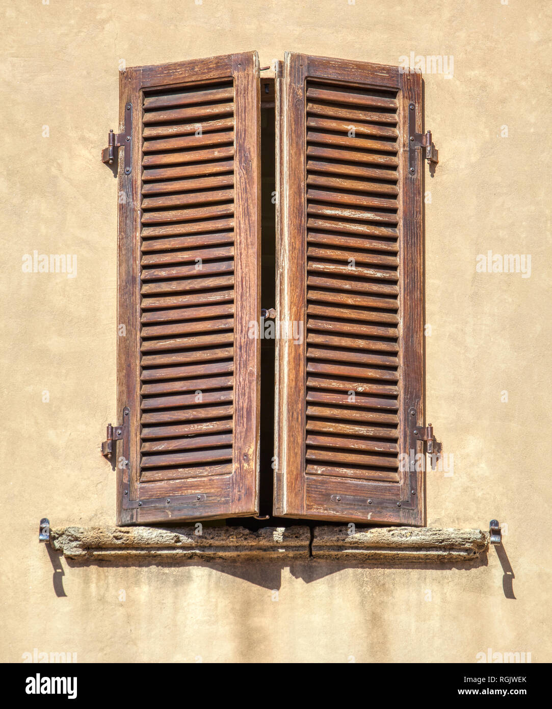 Photograph of a weathered, wood brown window shutter with black wrought iron hinges against a faded yellow plaster wall. Stock Photo