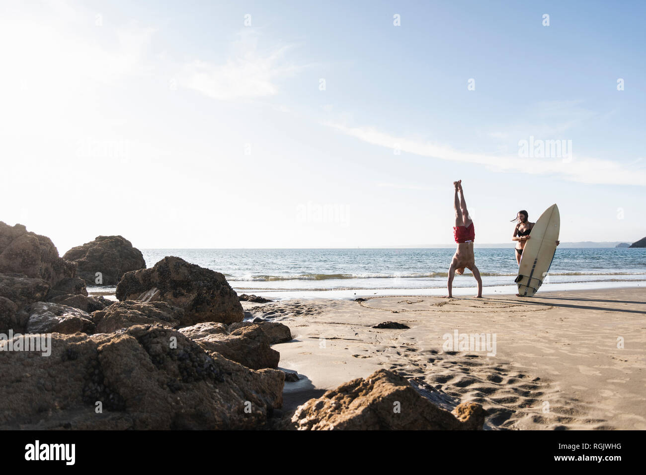 France, Brittany, young man doing a handstand and woman holding surfboard on the beach Stock Photo