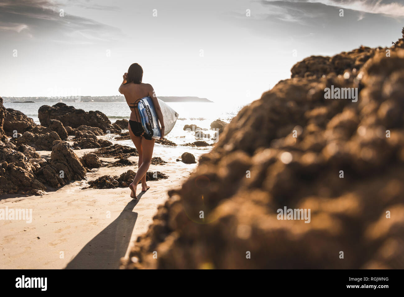 France, Brittany, young woman carrying surfboard on a rocky beach at the sea Stock Photo