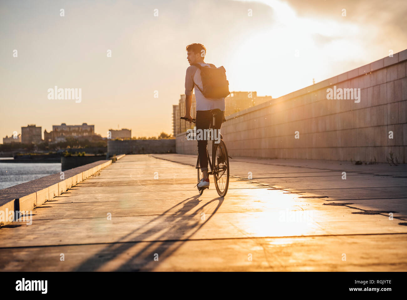 Young man with backpack riding bike on waterfront promenade at the riverside at sunset Stock Photo