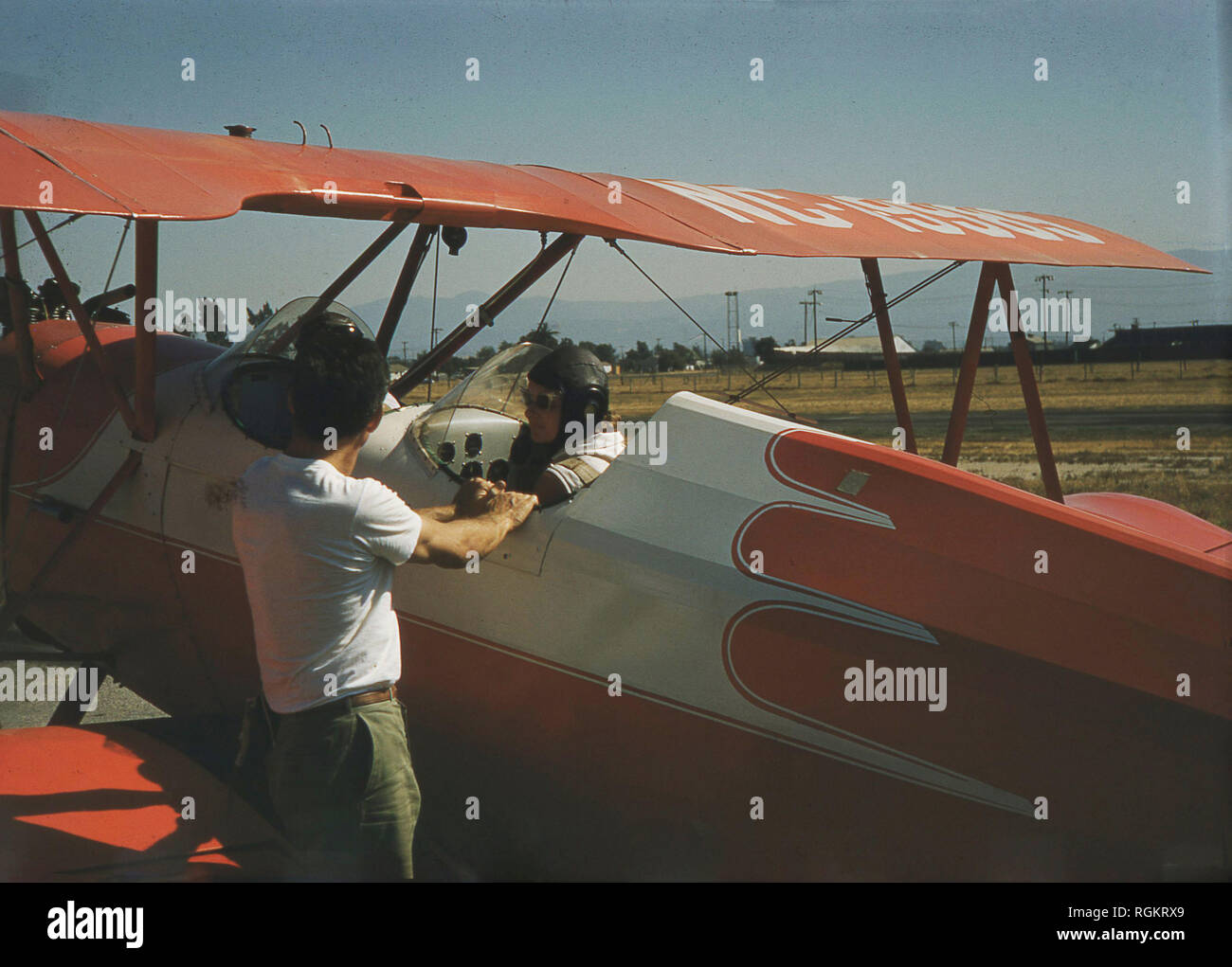 1950s, a female pilot wearing a leather flying hat and sitting in the second seat of a two-seater red coloured biplane having a conversation on the runway before take-off, USA. Biplanes or Fixed-wing aircraft are of historical importance in the history of aviation, as the first powered, controlled aeroplane to fly, the 'Wright Flyer' used a biplane wing system. Stock Photo