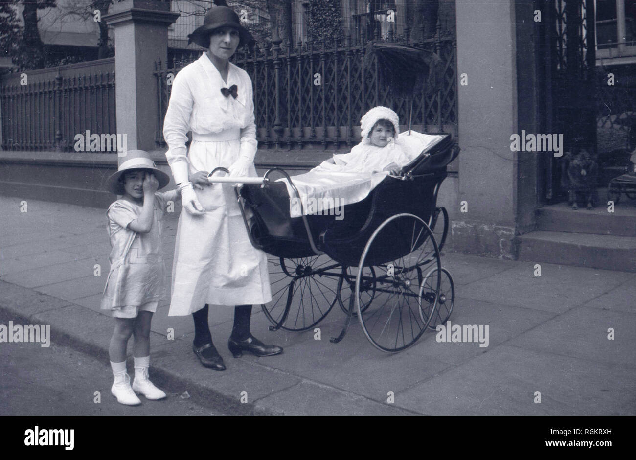 1920s, historical, on a pavement outside in a street, an elegant lady in dress of the day, possibly a nanny given her white dress, with a young girl and infant child sitting in a traditional coachbuilt baby carriage or pram, England, UK. With its large back wheels and suspension, this pram was comfortable for both the child riding in it and the mother or nanny pushing it. Stock Photo
