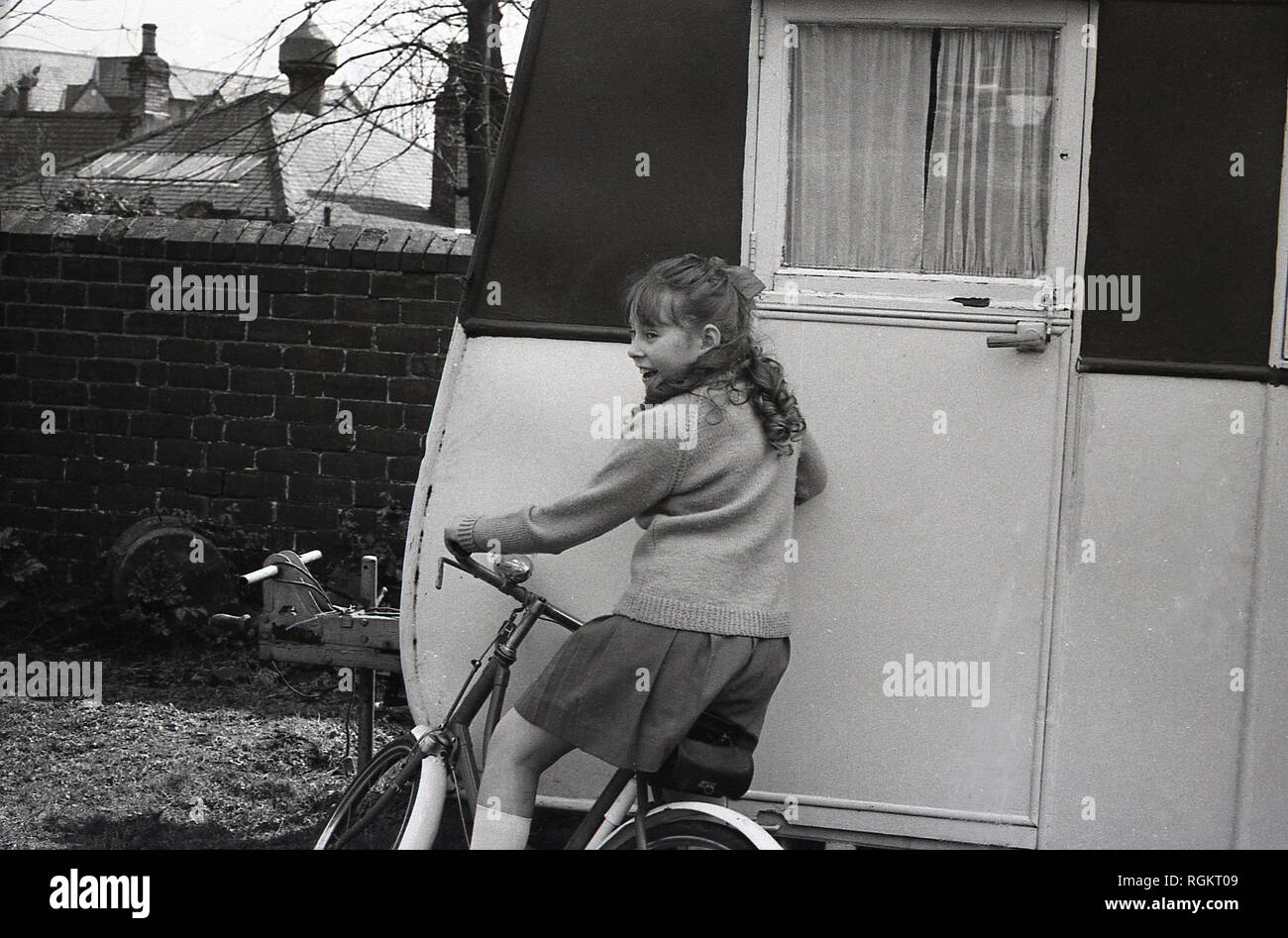 1960s, young schoolgirl on a bicycle by old caravan in a garden. Stock Photo