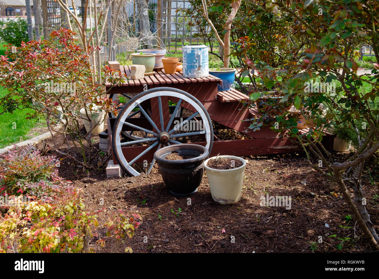 Rustic red wooden picnic table outside  in garden with an old wagon wheel next to it & various flower pots on the table and benches. Trees & bushes. Stock Photo