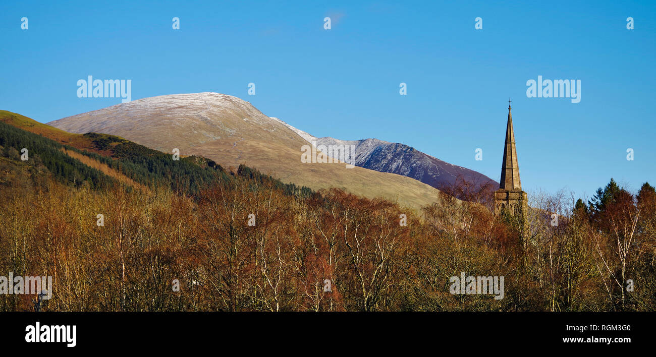 Tall spire of Keswick St John Church seen above woodland, Blease Fell and Blencathra summit ridge rising behind, winter, Lake District UK. Stock Photo