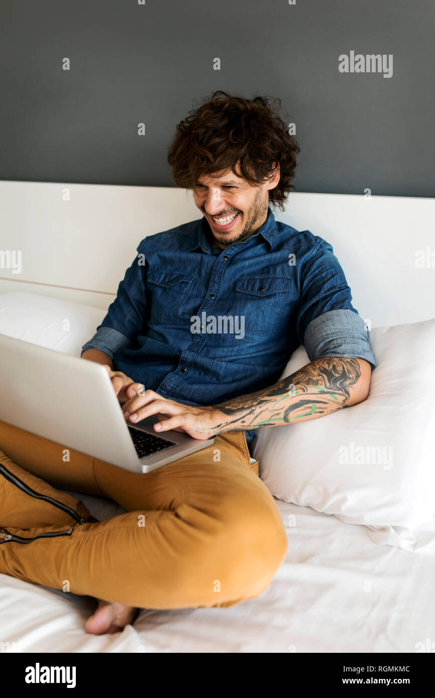 Happy tattooed man sitting on bed using laptop Stock Photo