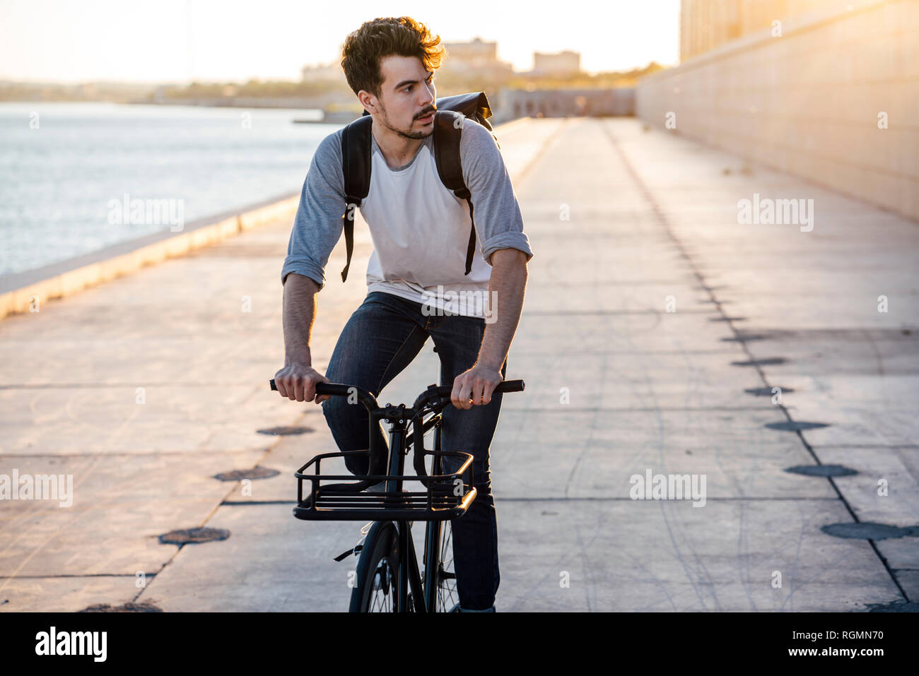 Young man with backpack riding bike on waterfront promenade at the riverside Stock Photo