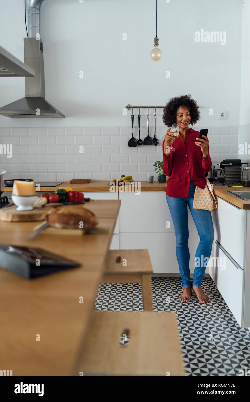 Woman drinking white wine in her kitchen, using smartphone Stock Photo