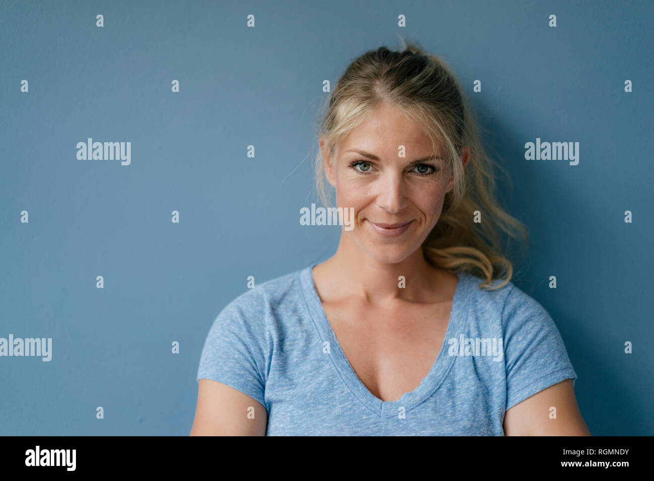 Portrait of smiling blond woman standing at blue wall Stock Photo