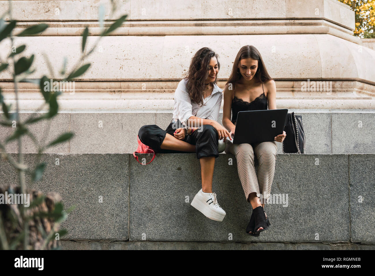 Two friends sitting side by side on a wall using laptop Stock Photo