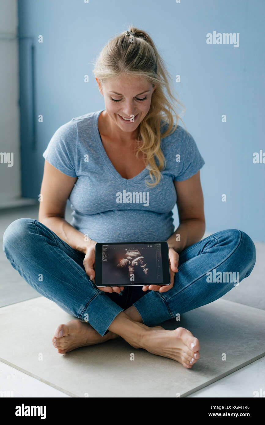 Smiling pregnant woman sitting on the floor showing ultrasound picture on tablet Stock Photo