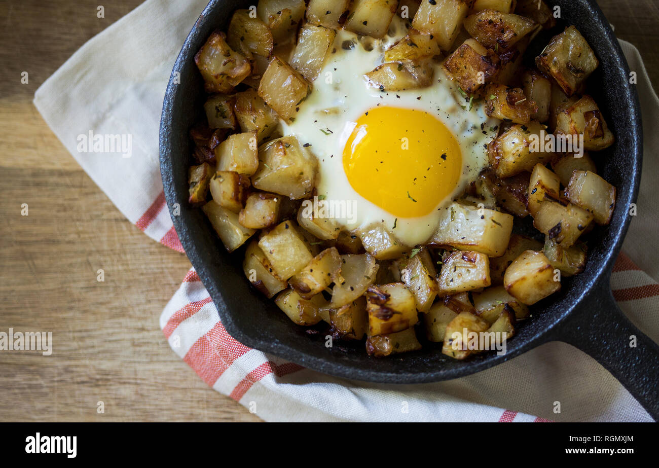 cast iron skillet with potato hash and fried egg on rustic wood surface with linen towel. Stock Photo