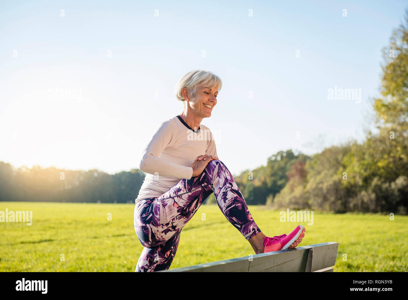 Smiling senior woman stretching on a bench in rural landscape Stock Photo