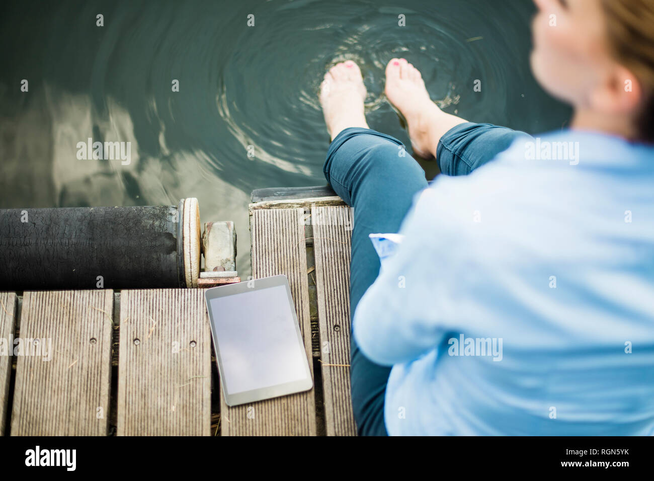 Woman sitting on jetty at a lake next to tablet with feet in water Stock Photo