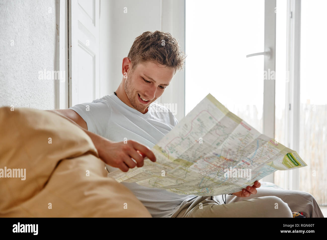 Smiling young man sitting on couch at home reading map Stock Photo