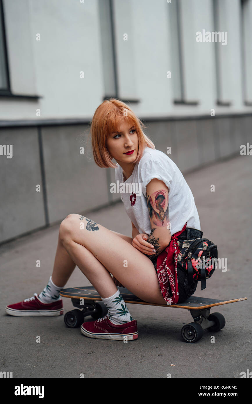 Young woman sitting on carver skateboard on the sidewalk Stock Photo