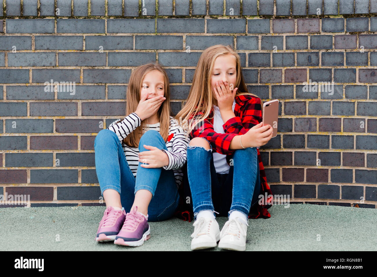 Portrait of two girls sitting in front of brick wall taking selfie with smartphone Stock Photo