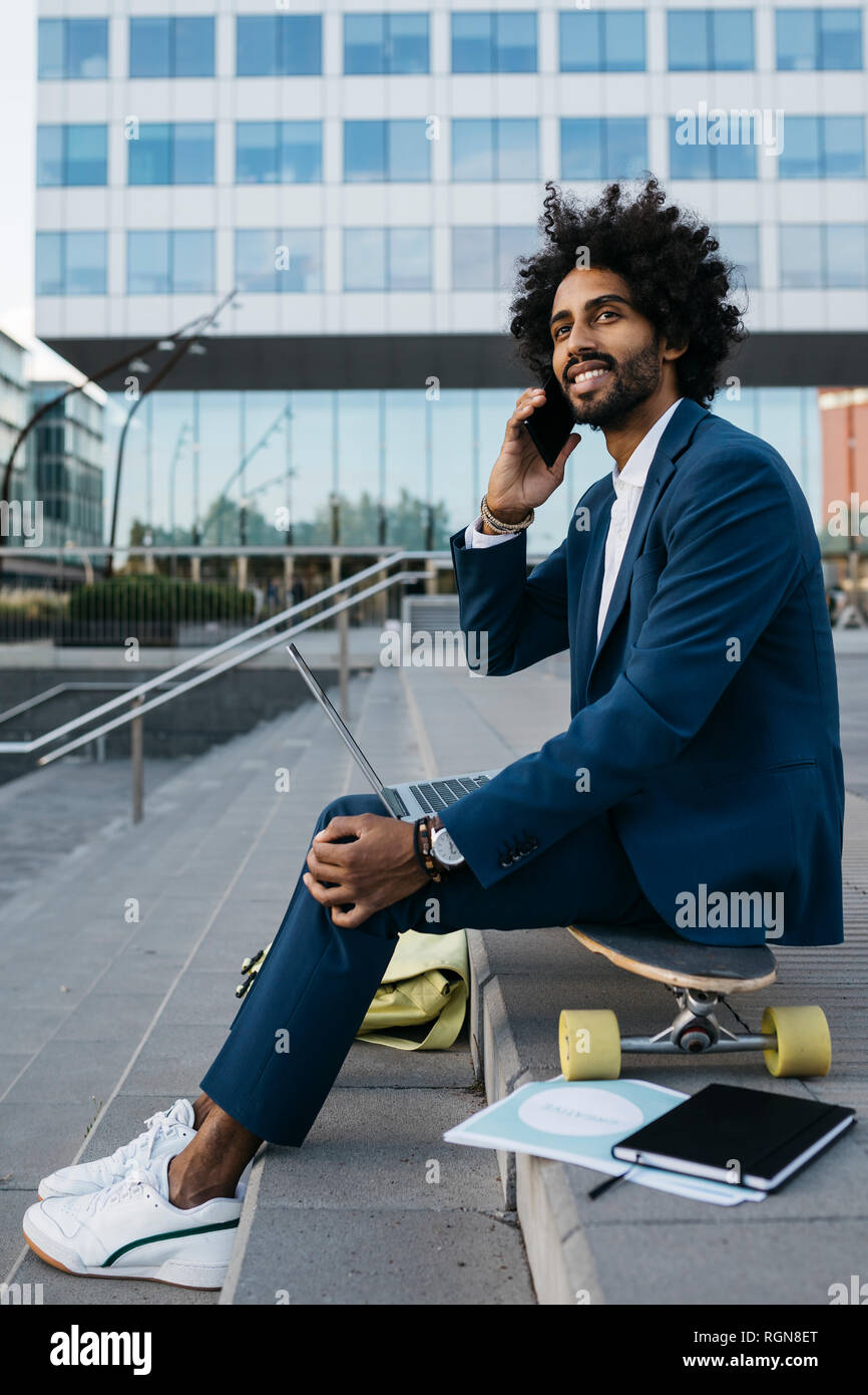 Spain, Barcelona, young businessman sitting outdoors in the city using cell phone and laptop Stock Photo