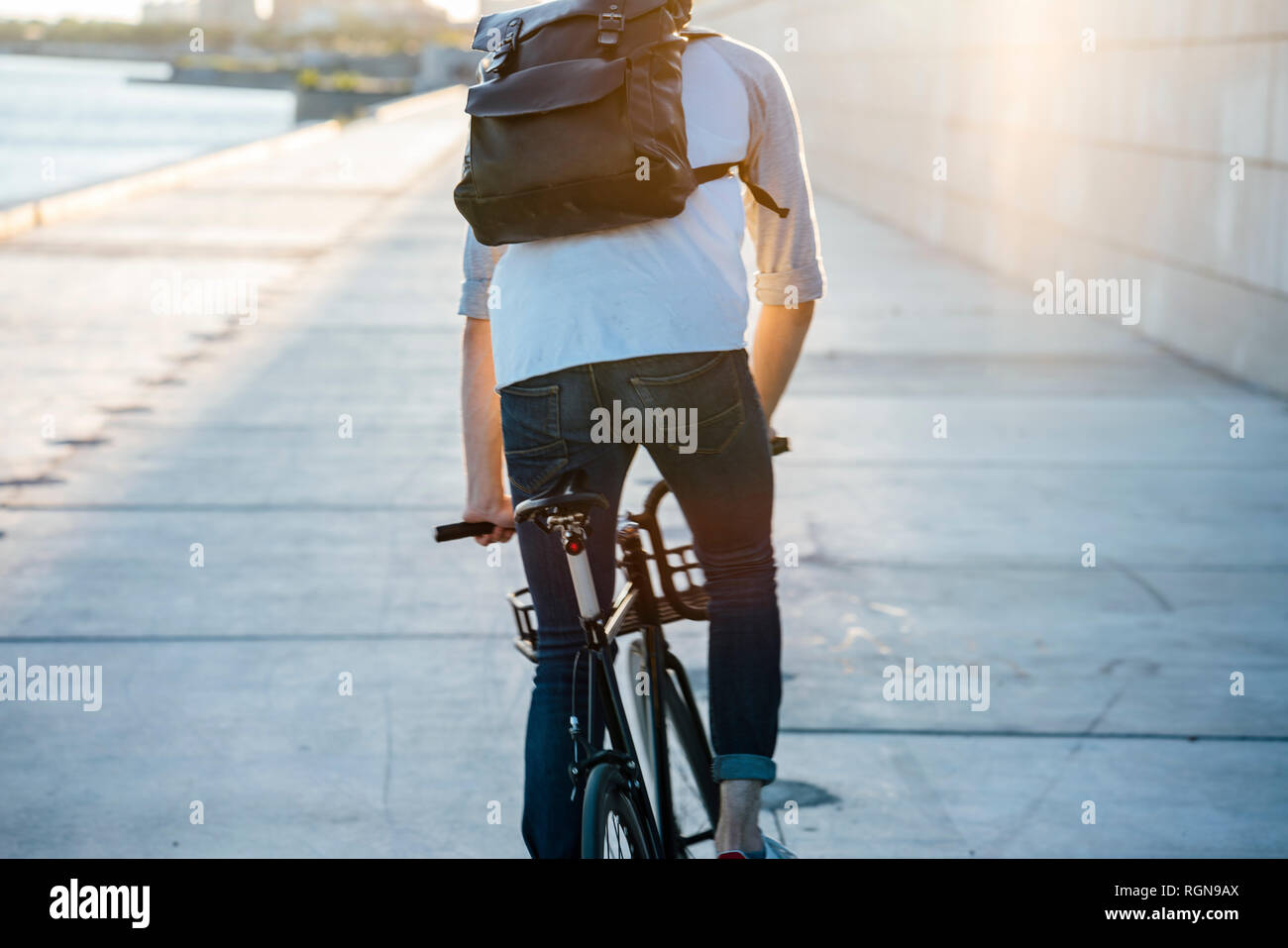 Rear view of young man with backpack riding bike on waterfront promenade at the riverside at