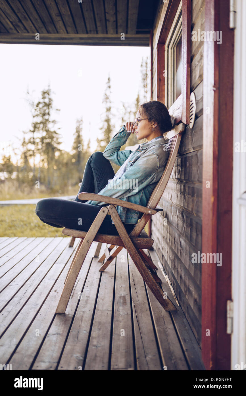Sweden, Lapland, pensive young woman sitting on chair on veranda looking at distance Stock Photo