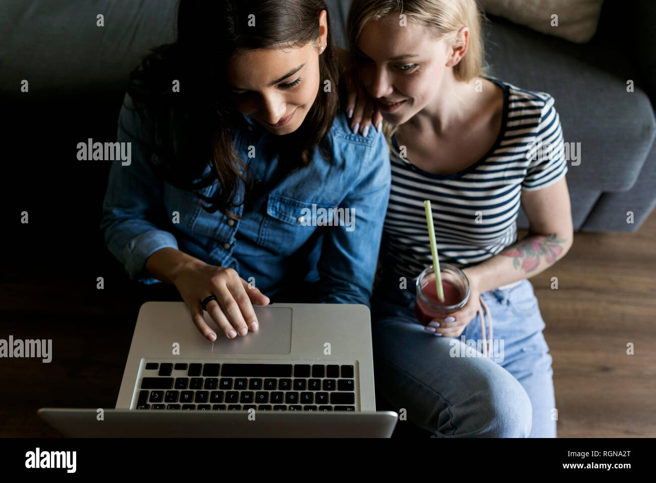 Two smiling young women sitting on floor with soft drink sharing laptop Stock Photo
