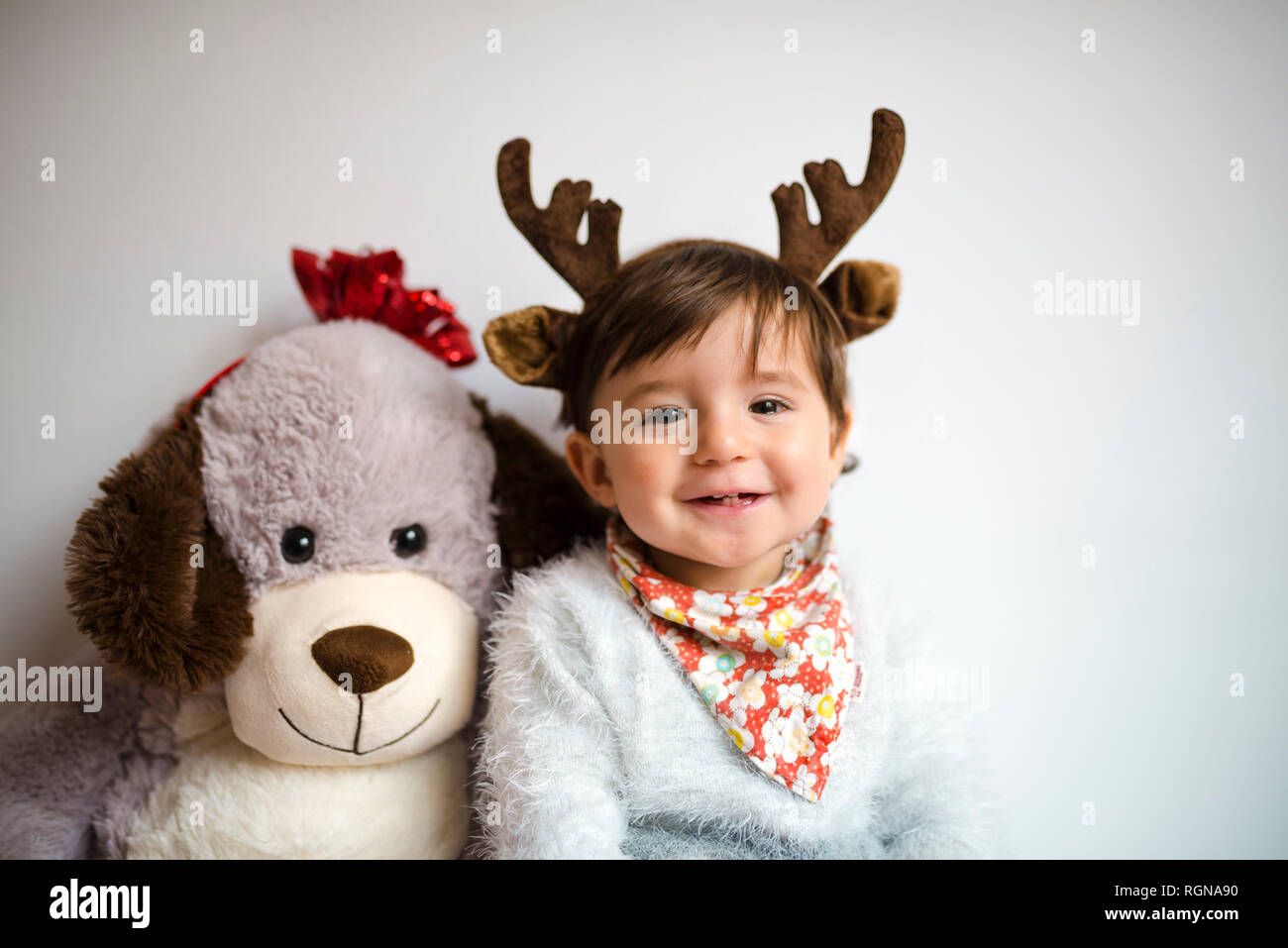 Portrait of happy baby girl with reindeer antlers headband beside her toy dog Stock Photo