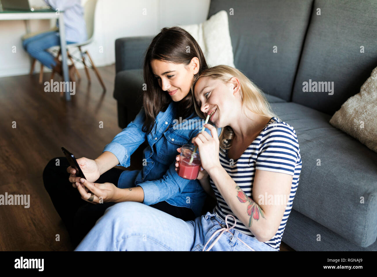 Two smiling young women sitting on floor with soft drink sharing cell phone Stock Photo