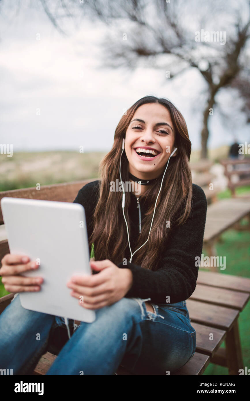 Portrait of laughing young woman sitting on bench outdoors using tablet and earphones Stock Photo