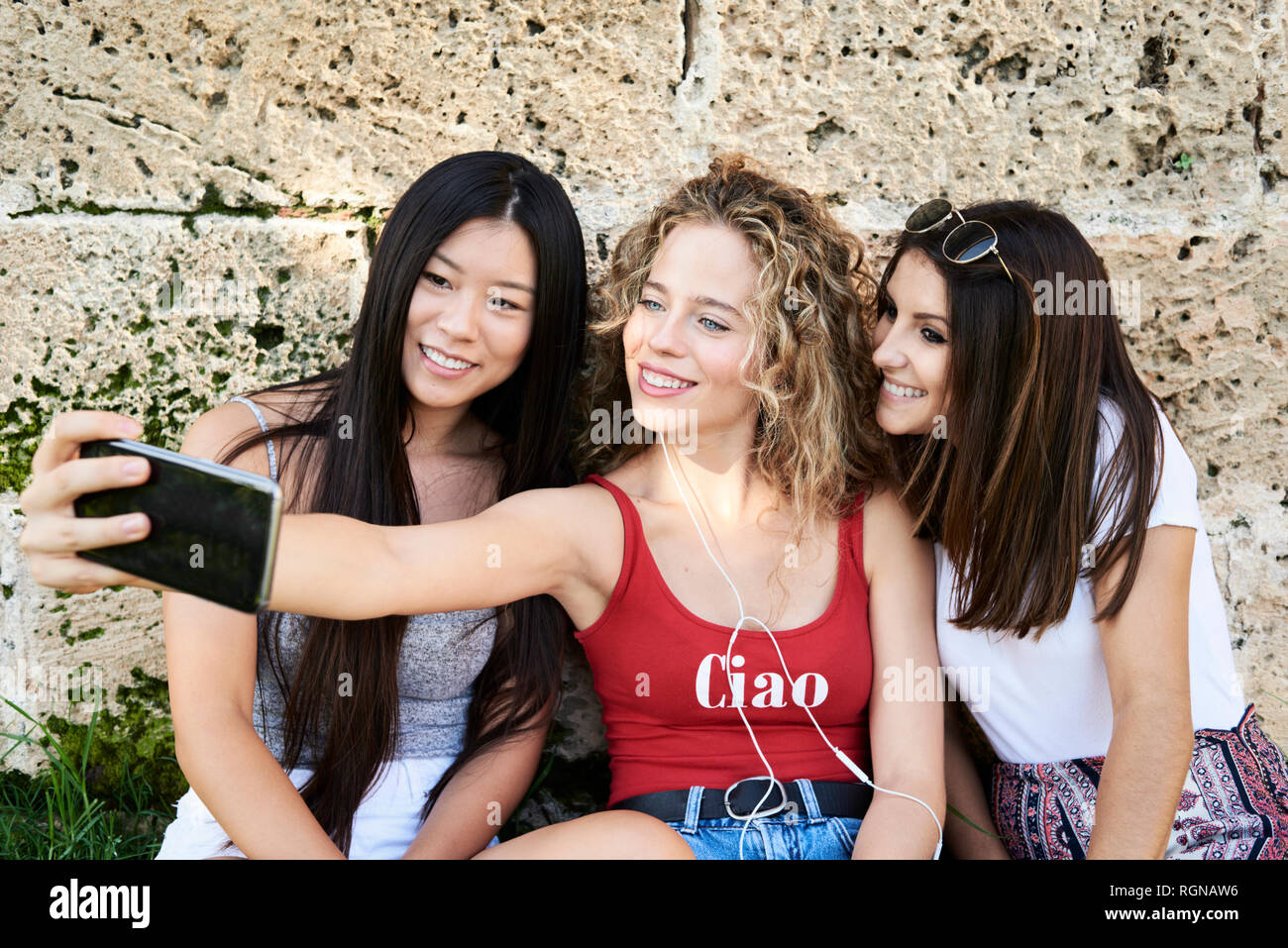 Three smiling young women sitting at stone wall taking a selfie Stock Photo