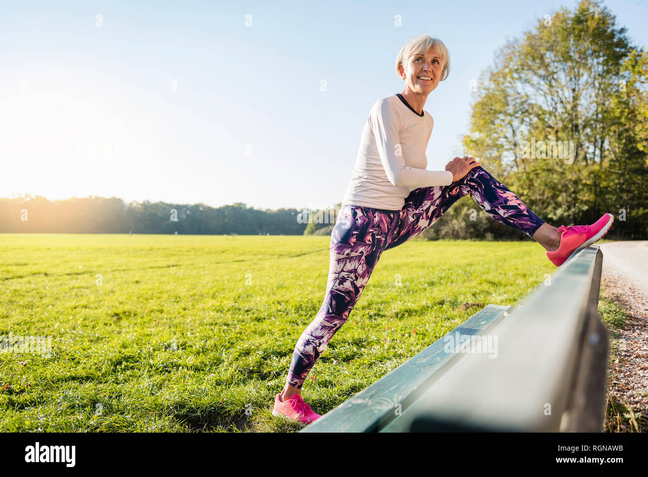 Smiling senior woman stretching on a bench in rural landscape Stock Photo