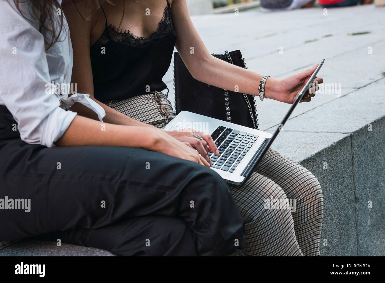 Two friends sitting side by side on a wall using laptop, partial view Stock Photo