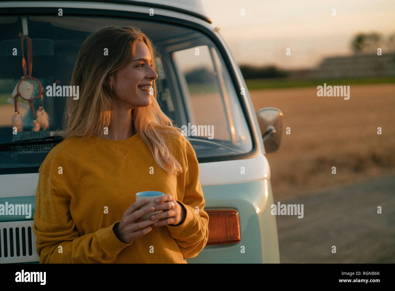 Happy young woman holding mug at camper van in rural landscape at sunset Stock Photo