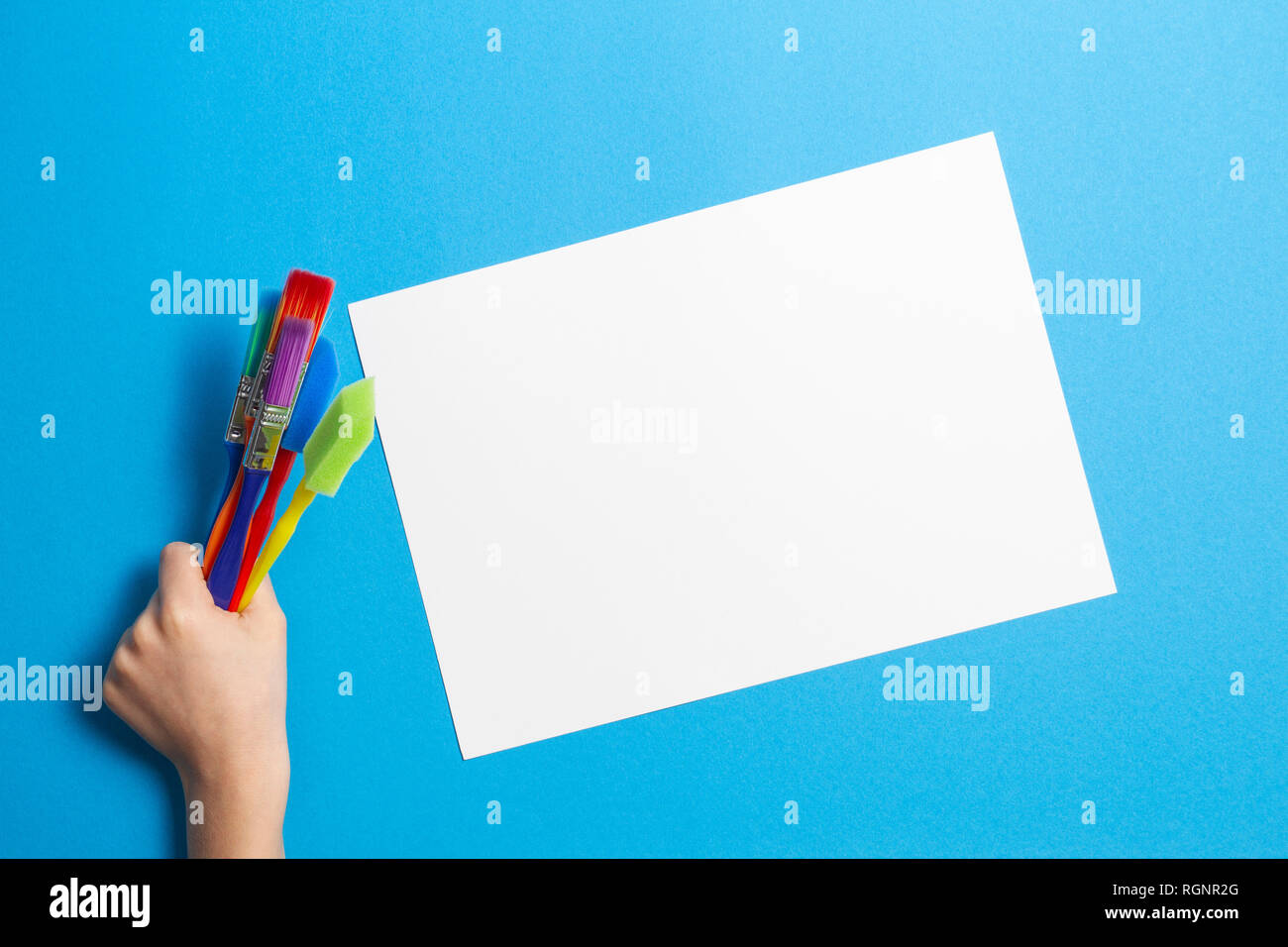 Child hand with colorful paintbrushes and blank white paper sheet on blue background Stock Photo