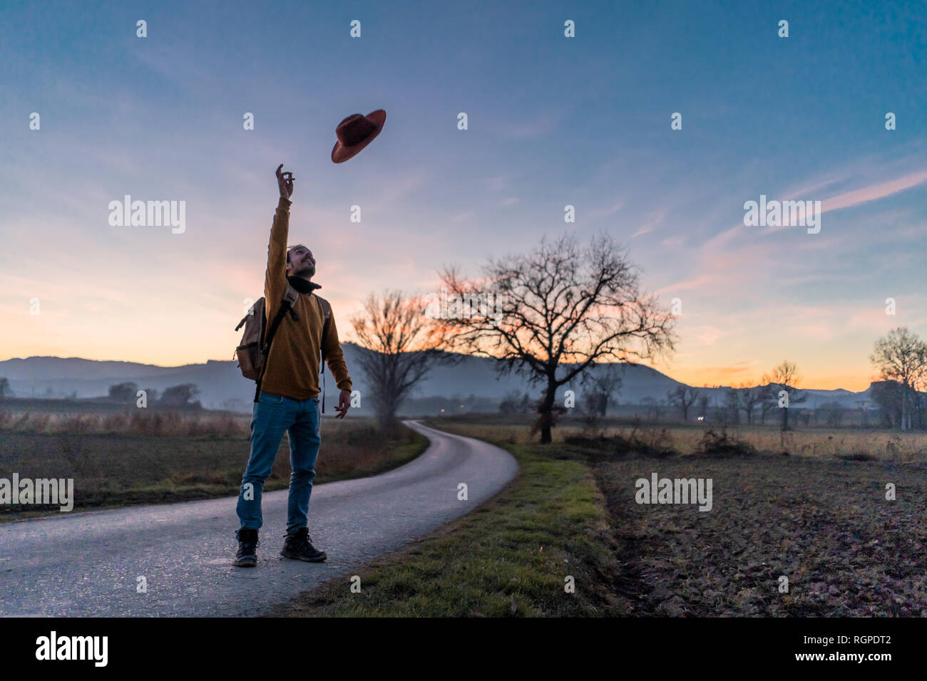 Adult male with backpack throwing stylish hat in air while standing on  narrow asphalt road in evening in majestic Spanish countryside Stock Photo  - Alamy