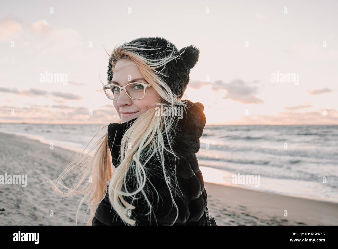 Back view of smiling blond attractive lady in eyeglasses and fur coat walking on coast near water at sunset in Klaipeda, Lithuania Stock Photo