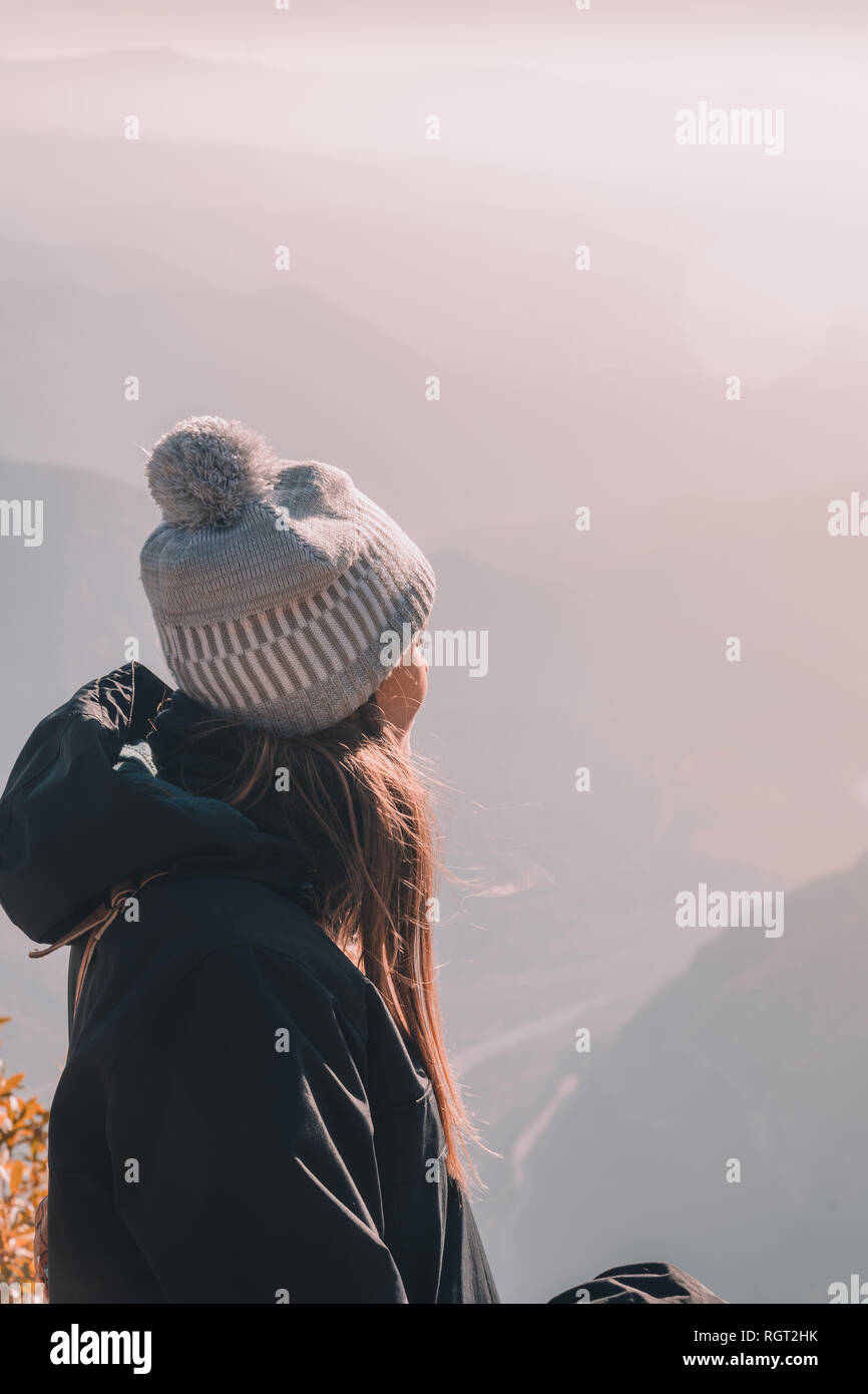 Side view of young lady in coat and hat looking on valley from top of mountain and cloudy sky Stock Photo