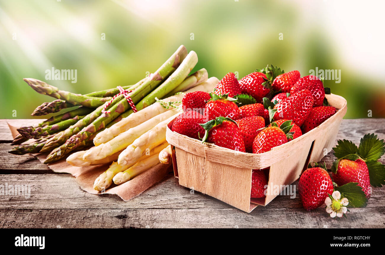 Wooden punnet of strawberries with fresh white and green asparagus shoots tied in bundles on an outdoor table in the garden or at farmers market Stock Photo