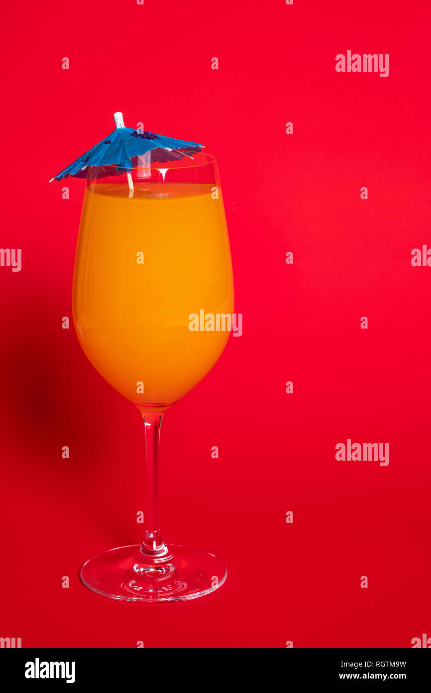Orange drink with a blue umbrella in a wine glass set against a solid red background. Stock Photo