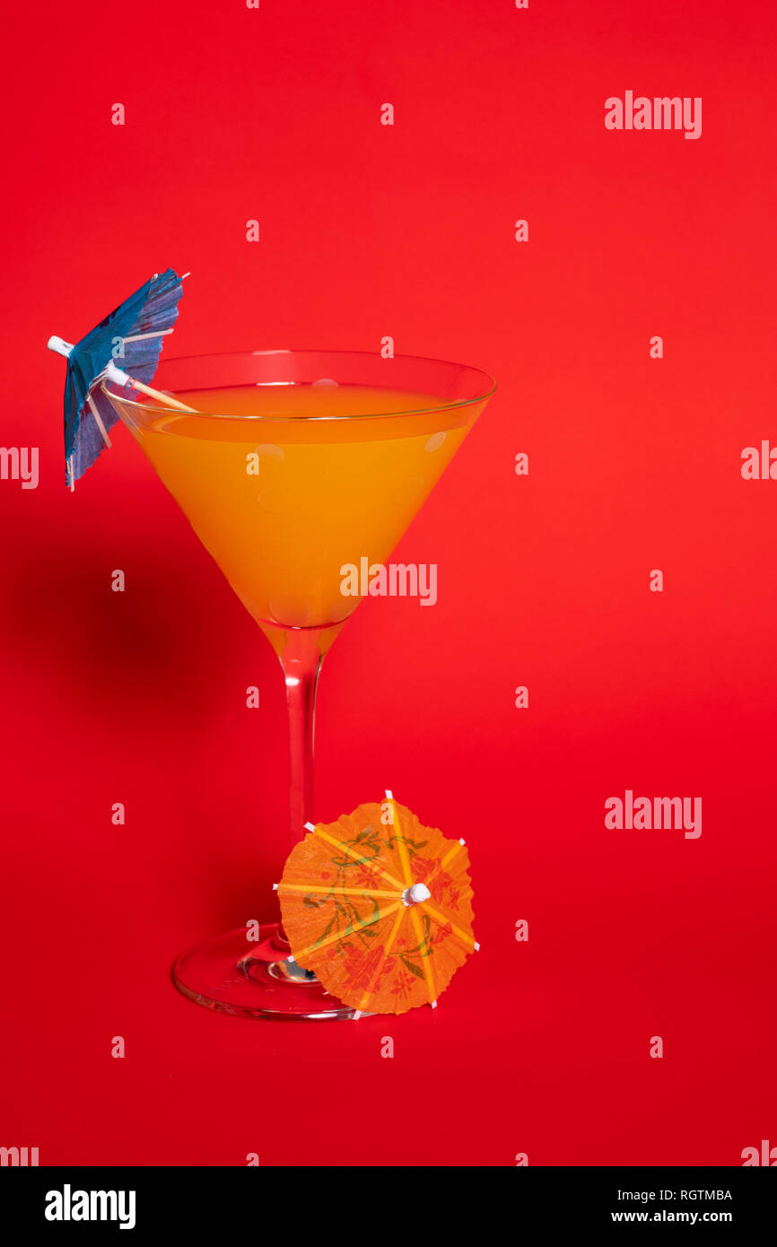 Orange drink with a blue umbrella in a martini glass set against a solid red background. An orange umbrella lies at the base of the glass. Stock Photo