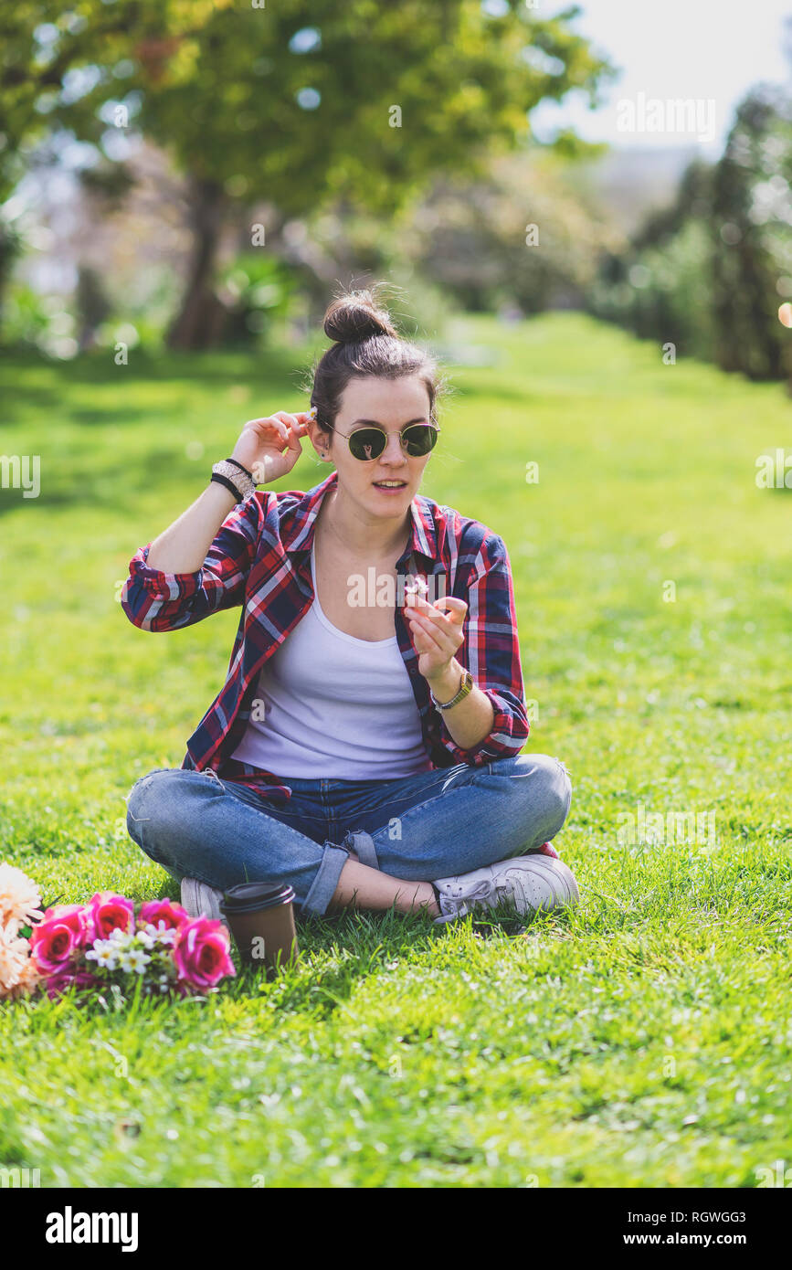 Front view of a young hipster Woman sitting on grass in a park while ...
