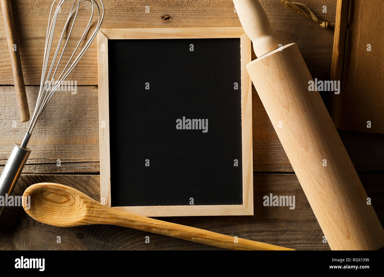 Blank, empty, black chalkboard with wooden rolling pin, wire whisk and recipe book flat lay from above on brown wooden table with copy space Stock Photo