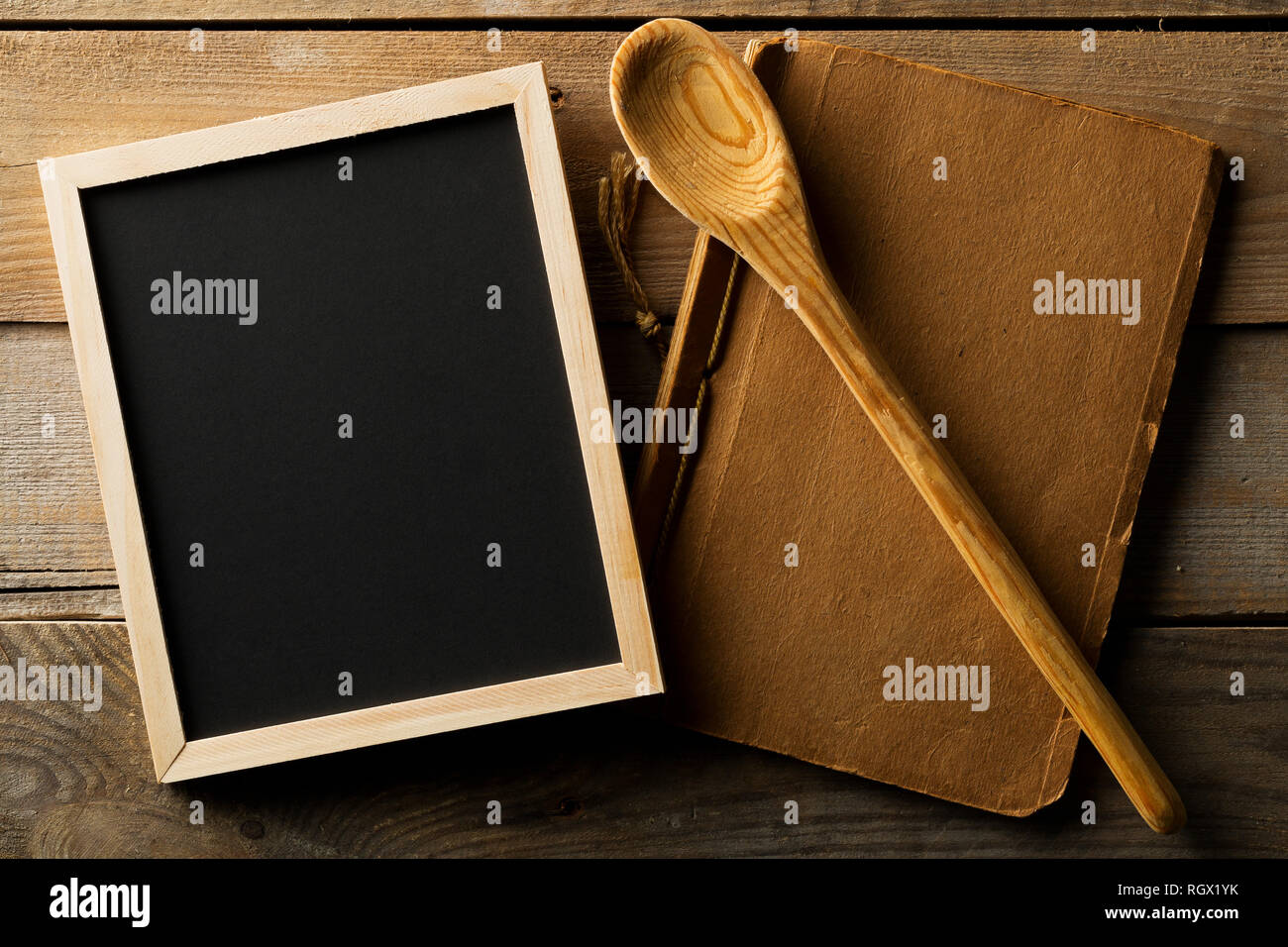 Blank, empty, black chalkboard with wooden cooking spoon and old recipe book flat lay from above on black wooden table with copy space Stock Photo