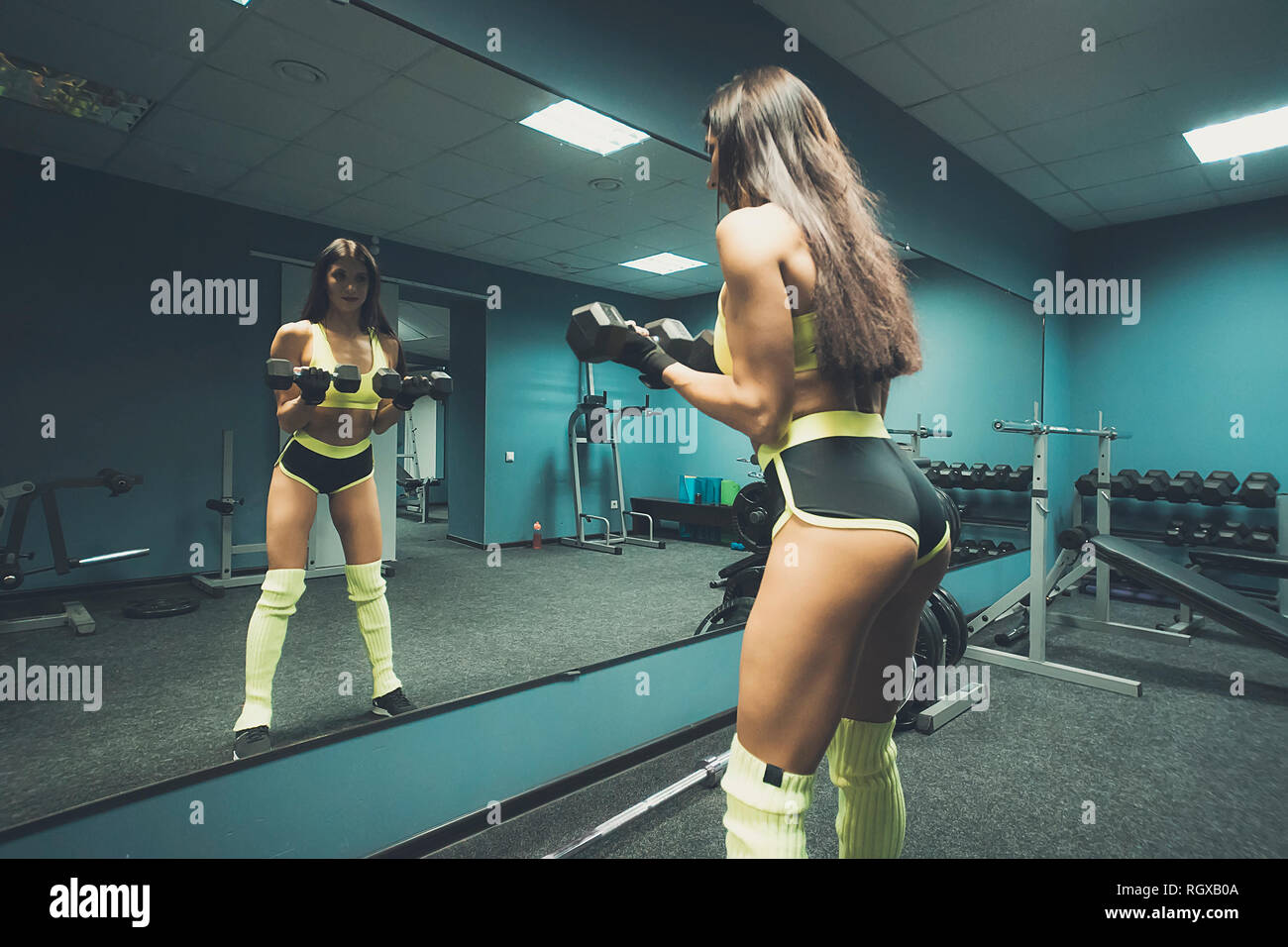 Female fitness coach with long dark hair in top and tiny sport shorts posing with dumbbells against the mirror of empty gym room. Sporty woman lifting Stock Photo
