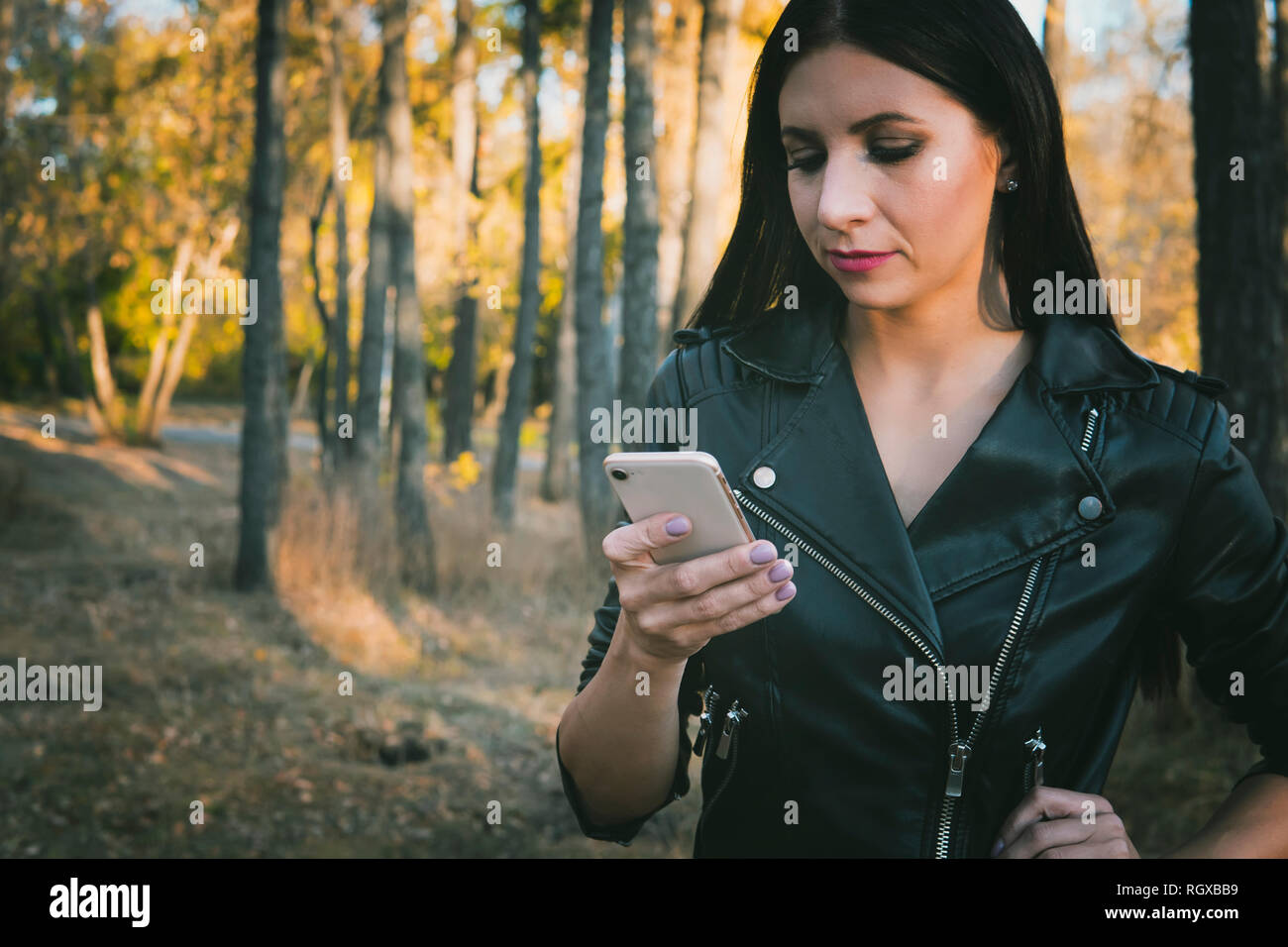 Autumn walk. Technologies. Pretty girl in leather clothes is using smartphone while walking in the park. Girl stays online being in autumn park. Woman Stock Photo