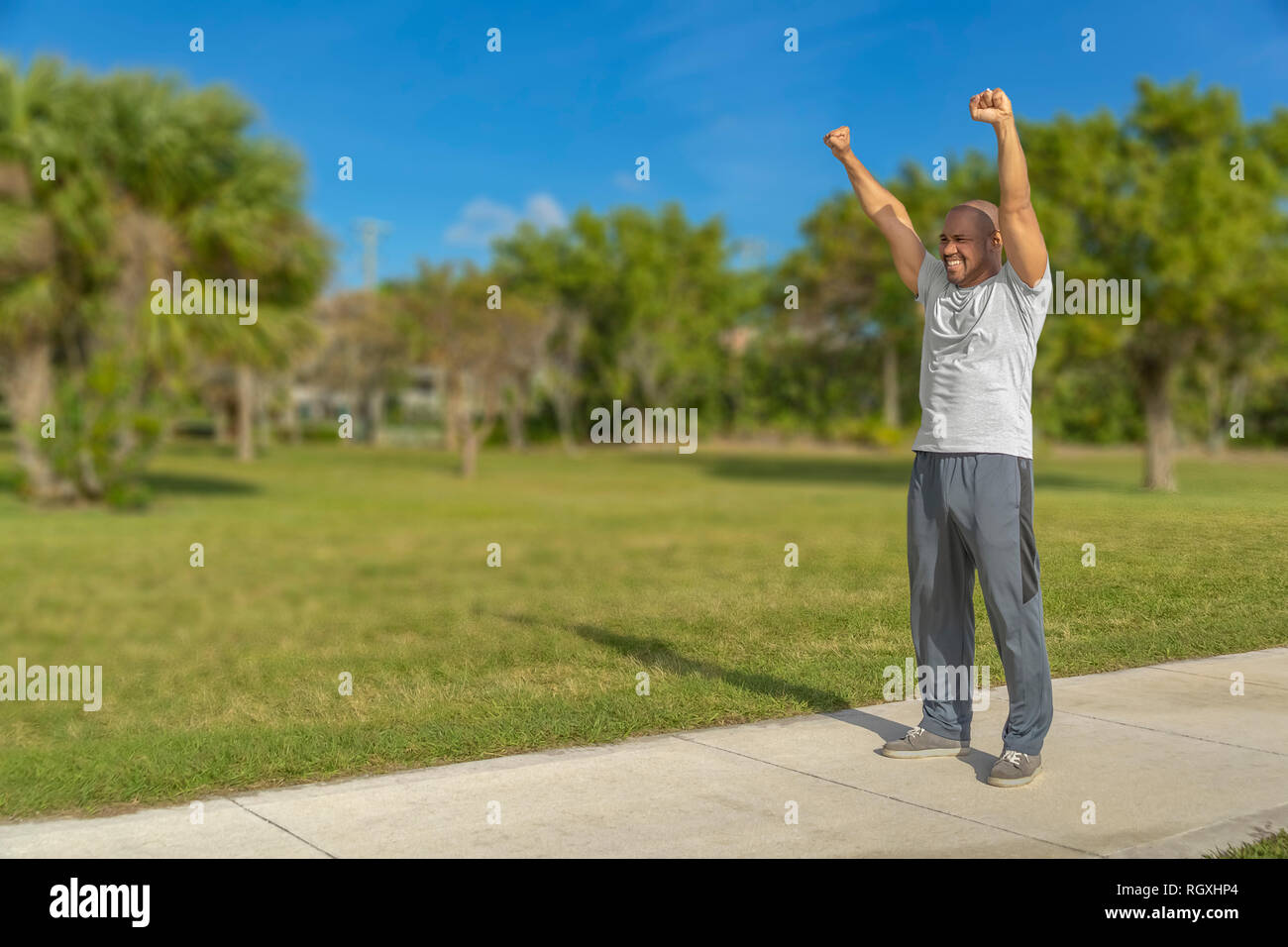 A black man shows his victory excitement by stretching his arms up high with a smile standing on the running path at the park. Stock Photo