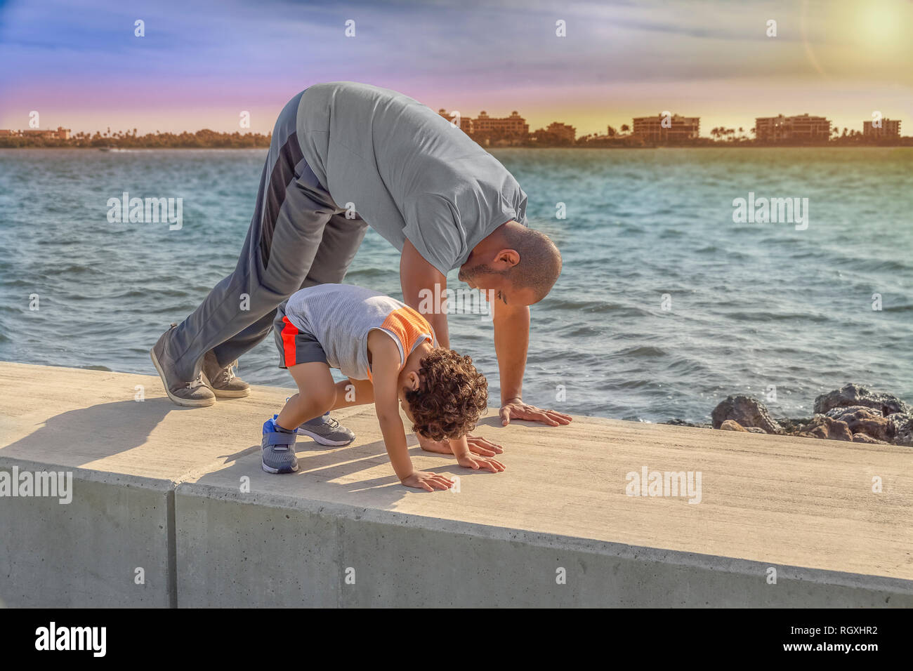 Father and young son start the morning exercise with stretches on the waterfront wall ledge as the sun rises. Stock Photo