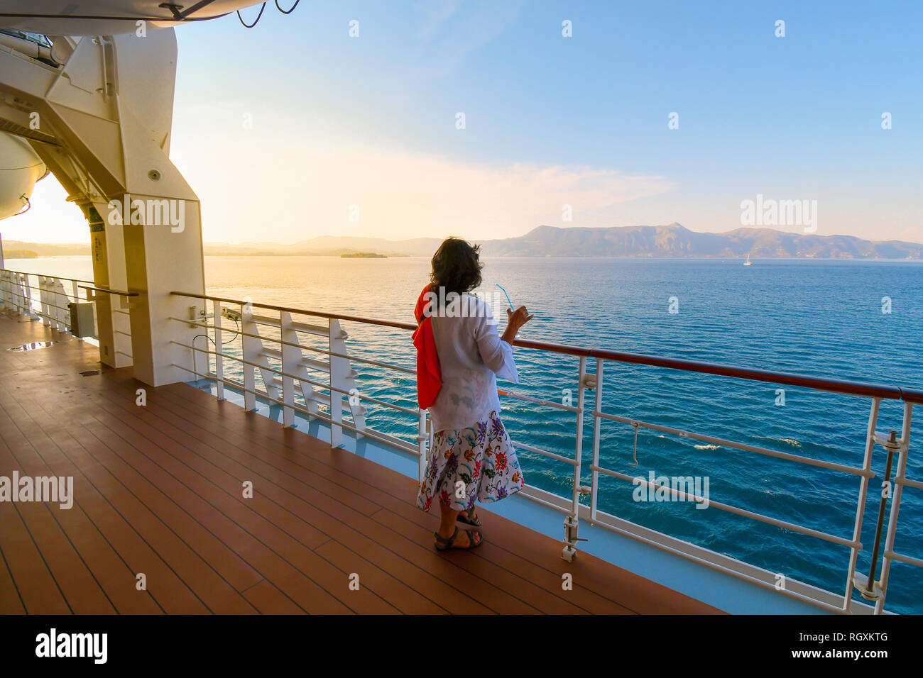 A woman sips a drink on the deck of a cruise ship as the sun sets and the ship passes islands on the Aegean Sea Stock Photo