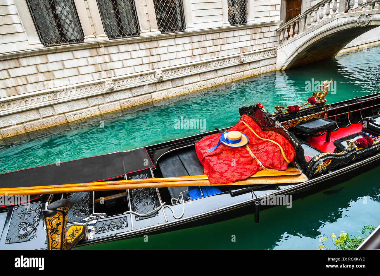 A fish swims under Venetian gondola with oars, gondolier hat, striped shirt and red blanket as it sits unoccupied in a colorful canal in Venice Italy Stock Photo